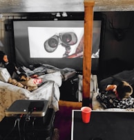 A cozy home theater setup with people lounging on couches and covered in blankets. They are watching a large screen with an animated robot character displayed. The space includes a projector on a table and dim ambient lighting, with a decorative deer head mounted on the wall.