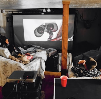 A cozy home theater setup with people lounging on couches and covered in blankets. They are watching a large screen with an animated robot character displayed. The space includes a projector on a table and dim ambient lighting, with a decorative deer head mounted on the wall.