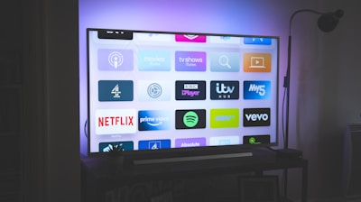 A flat-screen television is displayed in a dimly lit room, showcasing a variety of streaming service apps on its screen, such as Netflix, Prime Video, and Spotify. The TV is positioned on a black stand beside a tall floor lamp, casting a soft ambient light on the surrounding area.