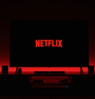 A television screen displays the Netflix logo in red against a black background. The room is dimly lit with a red glow possibly from LED backlighting behind the screen. Two speakers are positioned on either side of the TV set atop a minimalist entertainment center.
