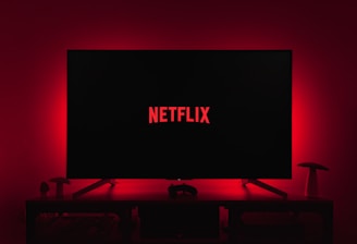 A dark room featuring a television screen displaying the word 'Netflix' in red against a black background. The screen is centered on a console table, with a video game controller placed in front. The room is dimly lit by red ambient lighting, creating a cozy atmosphere.