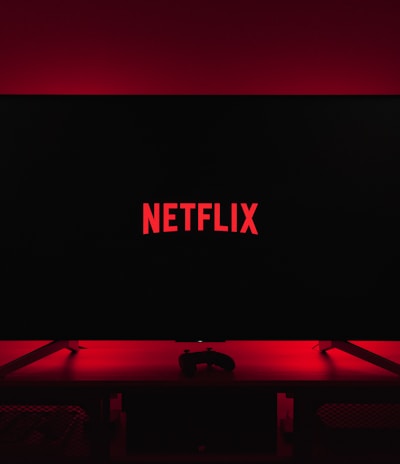 A dark room featuring a television screen displaying the word 'Netflix' in red against a black background. The screen is centered on a console table, with a video game controller placed in front. The room is dimly lit by red ambient lighting, creating a cozy atmosphere.