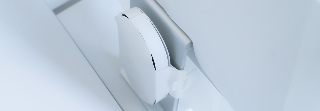 A white, sleek, vertical gaming console placed beside a large, slim monitor. The setting exudes minimalism with a focus on modern technology design.