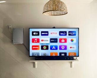 A modern living room features a wall-mounted flat-screen television displaying multiple streaming app icons, such as Apple TV, HBO Max, Netflix, Disney+, and Prime Video. Below the TV is a soundbar sitting on a small white shelf. A wicker pendant light hangs from the ceiling above, and the wall is painted a neutral color.