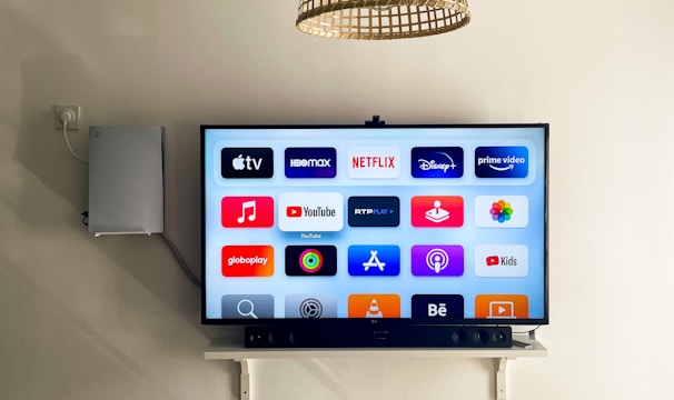 A modern living room features a wall-mounted flat-screen television displaying multiple streaming app icons, such as Apple TV, HBO Max, Netflix, Disney+, and Prime Video. Below the TV is a soundbar sitting on a small white shelf. A wicker pendant light hangs from the ceiling above, and the wall is painted a neutral color.