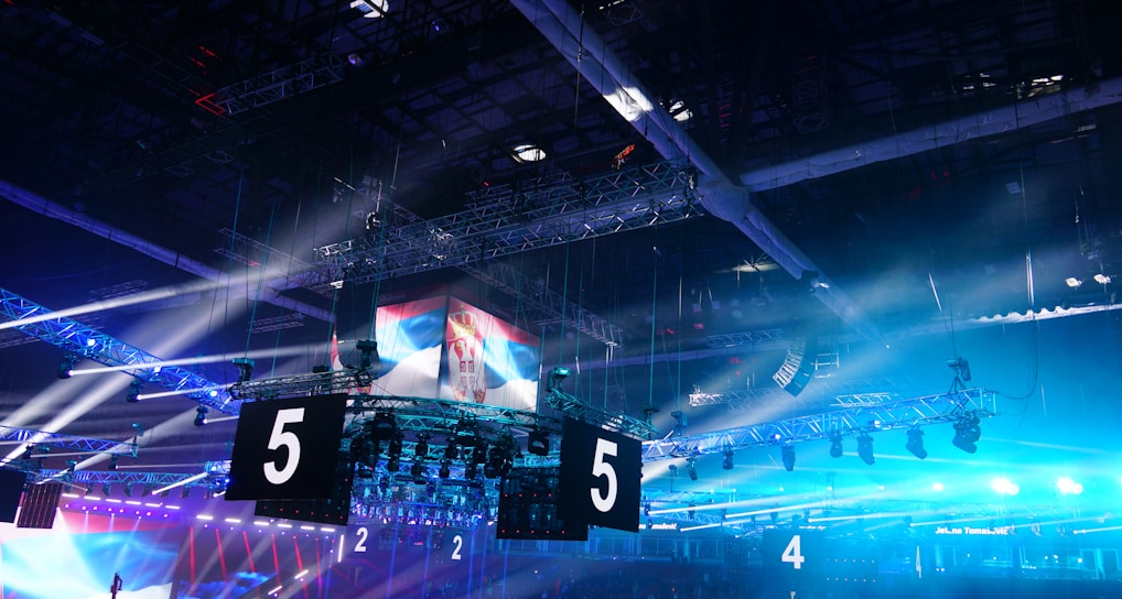 A large indoor arena is equipped with advanced lighting and audio systems. Suspended above the space are numerous digital displays showcasing numbers. The environment is filled with blue illumination and spotlights creating a vibrant atmosphere.
