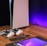 A sleek, modern setup featuring a gaming console with two controllers on a wooden surface. Nearby are a smartphone, smartwatch, tablet with a vivid screen, wireless earbuds, and a stylus. The background is illuminated with a soft purple glow.