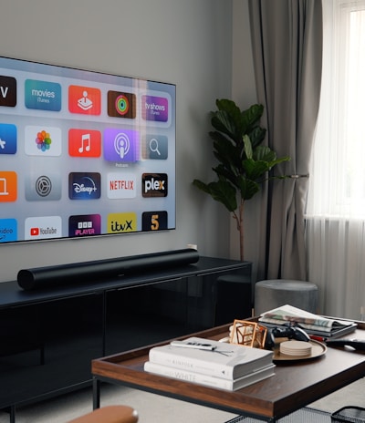 A modern living room featuring a mounted flat-screen TV displaying various streaming service icons. The room includes a black entertainment console beneath the TV, a brown wooden coffee table with books and remote controls, a gray sofa, and a tall green plant near a large window with sheer white curtains.
