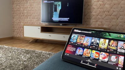 A modern living room features a flat-screen television mounted on a light-colored wooden stand with a patterned wall backdrop. Below the TV, a carpet lies on a wooden floor. In the foreground, a tablet displays a streaming platform interface with various show thumbnails, suggesting a relaxed entertainment setup.
