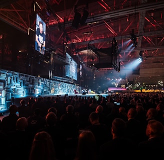 A large indoor event with a stage featuring bright screens and spotlights, attended by a substantial crowd. The venue has a high ceiling with industrial beams and red lighting accents. The atmosphere resembles a concert or a conference with a focus on visual effects.
